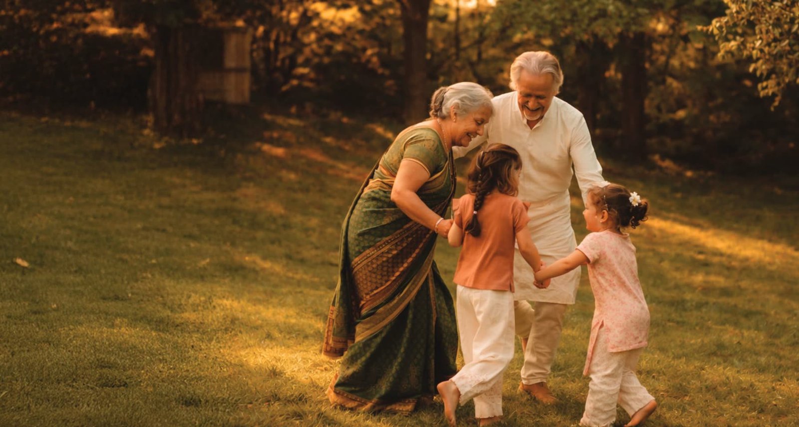 Grandparents dancing with their two granddaughters on a lawn in late golden light.
